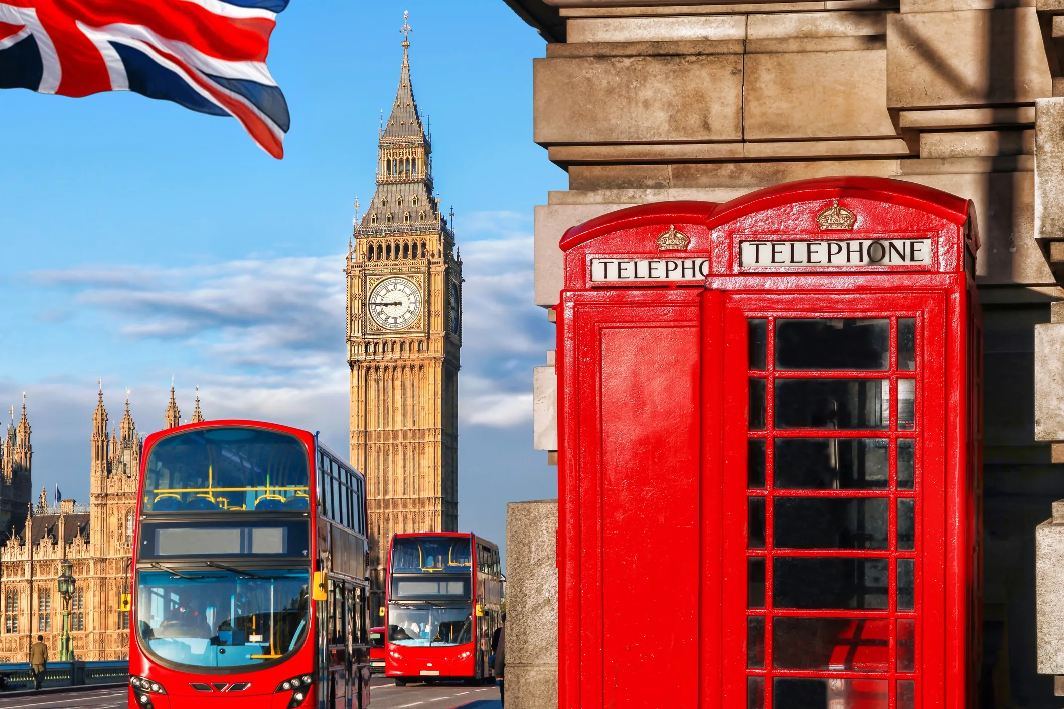 Photo of London with Big Ben in the background. Two red double-decker buses are driving down the street in front of it, and two red telephone boxes are standing a little further ahead.
